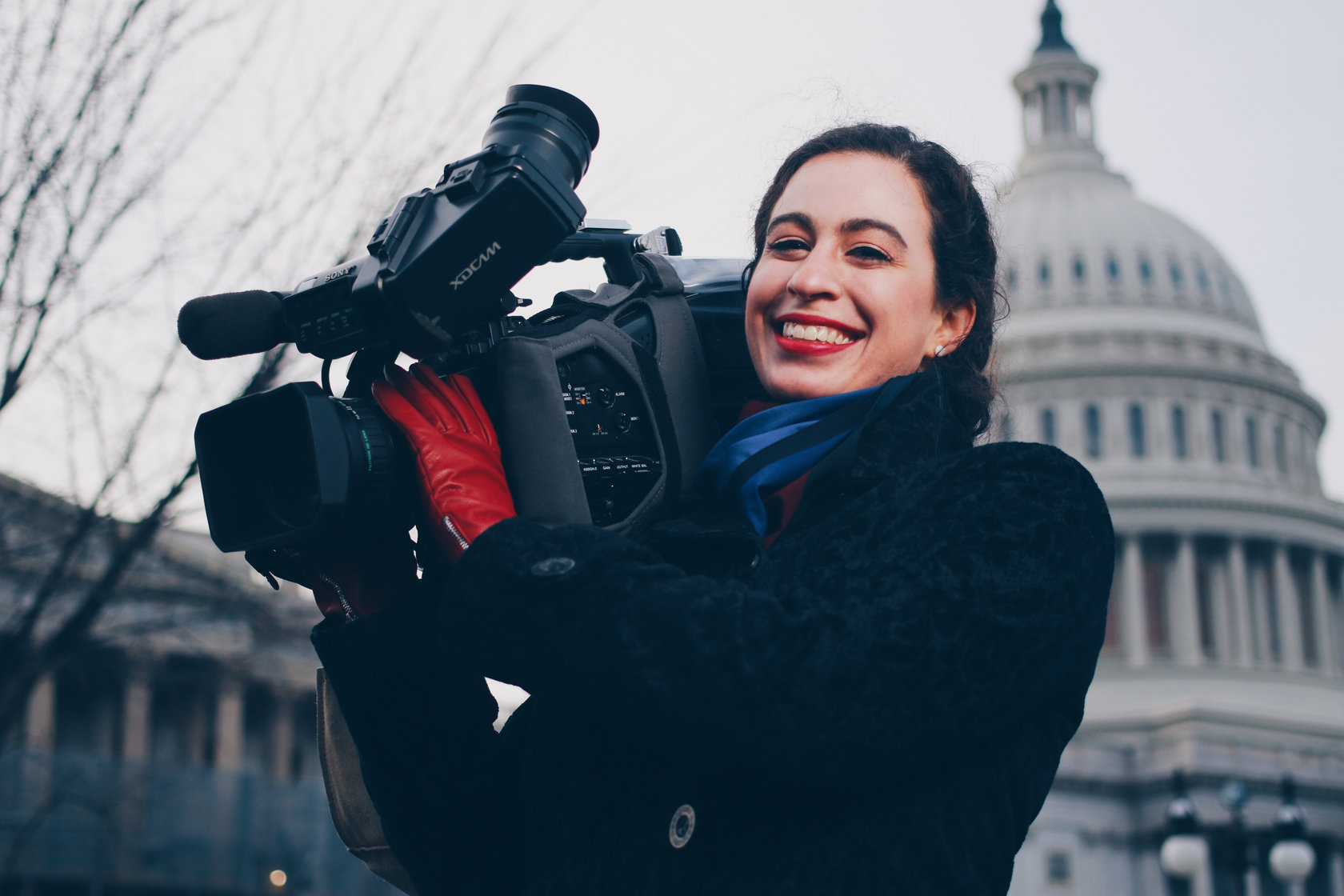 Maya Shwayder smiling with a broadcast camera on her shoulder in front of the U.S. Capitol